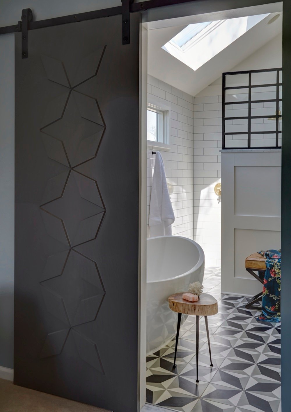 View through a geometric-paneled sliding door into the primary ensuite with freestanding tub, white subway tile, steel-framed shower screen and patterned cement floor, by Martire Custom Homes