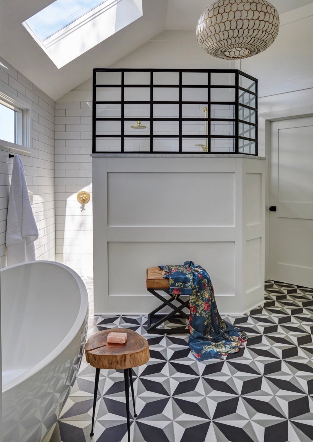 Primary ensuite with blackened-steel shower framework above a white paneled knee wall, freestanding tub, capiz shell pendant and geometric cement tile floor lit by a skylight, by Martire Custom Homes