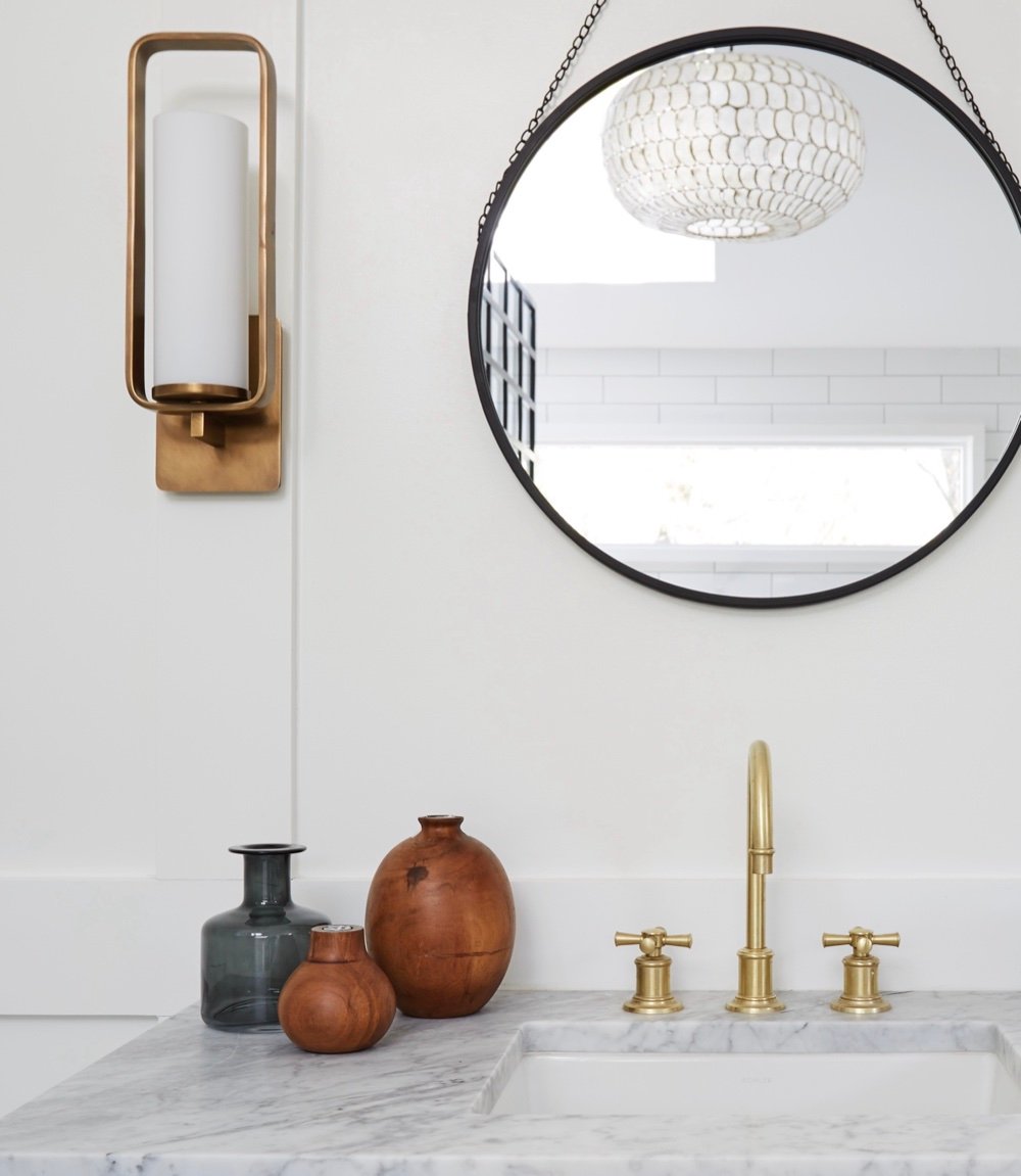 Bathroom vanity detail with a brass cylinder sconce, round black-framed mirror reflecting a capiz shell pendant, and brass cross-handle faucet on Carrara marble, by Martire Custom Homes
