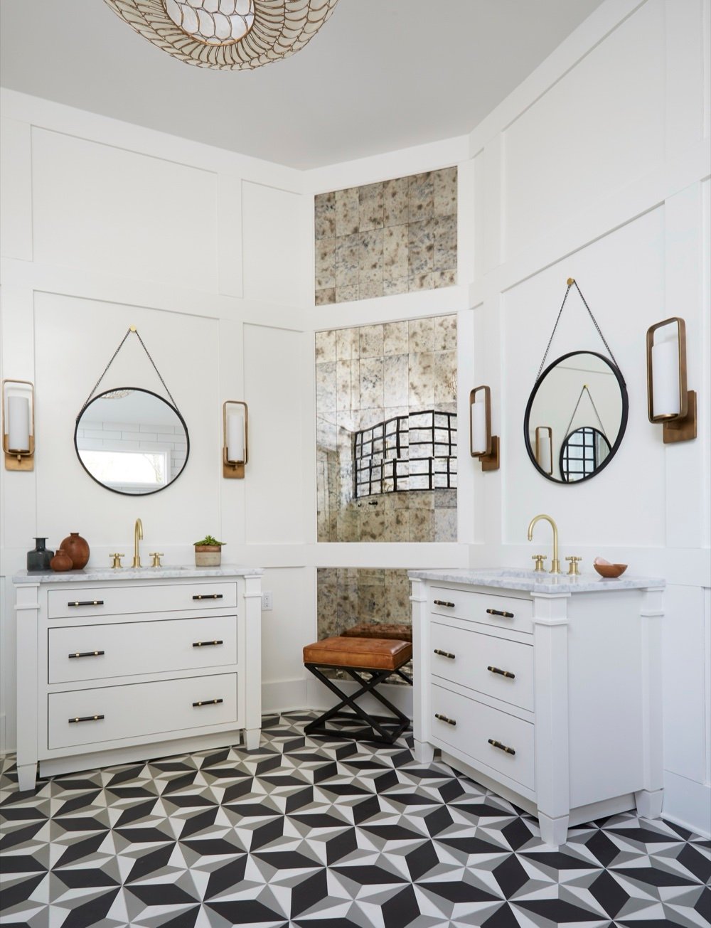 Primary ensuite with paired white vanities, black-and-brass hardware, brass sconces, a floor-to-ceiling antique mirror column and geometric cement tile floor, by Martire Custom Homes