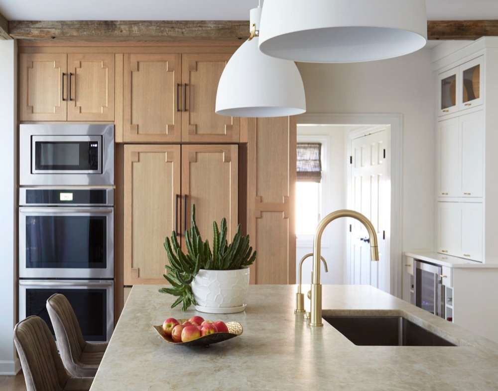 Kitchen wall of white oak cabinetry with applied geometric trim and black pulls concealing paneled refrigerator doors beside stainless double wall ovens, under a reclaimed wood beam, by Martire Custom Homes