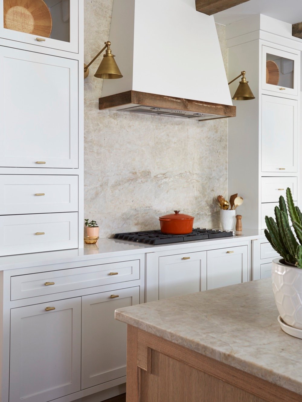 Kitchen range wall with full-slab cinnamon quartzite backsplash, white plaster hood trimmed in reclaimed wood, brass cone sconces and white cabinetry with brass pulls, by Martire Custom Homes
