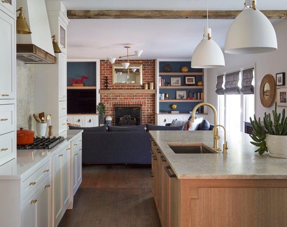 Open-plan kitchen with white oak island and cinnamon quartzite counters flowing into a family room with exposed brick fireplace, navy sectional and built-in shelving, by Martire Custom Homes