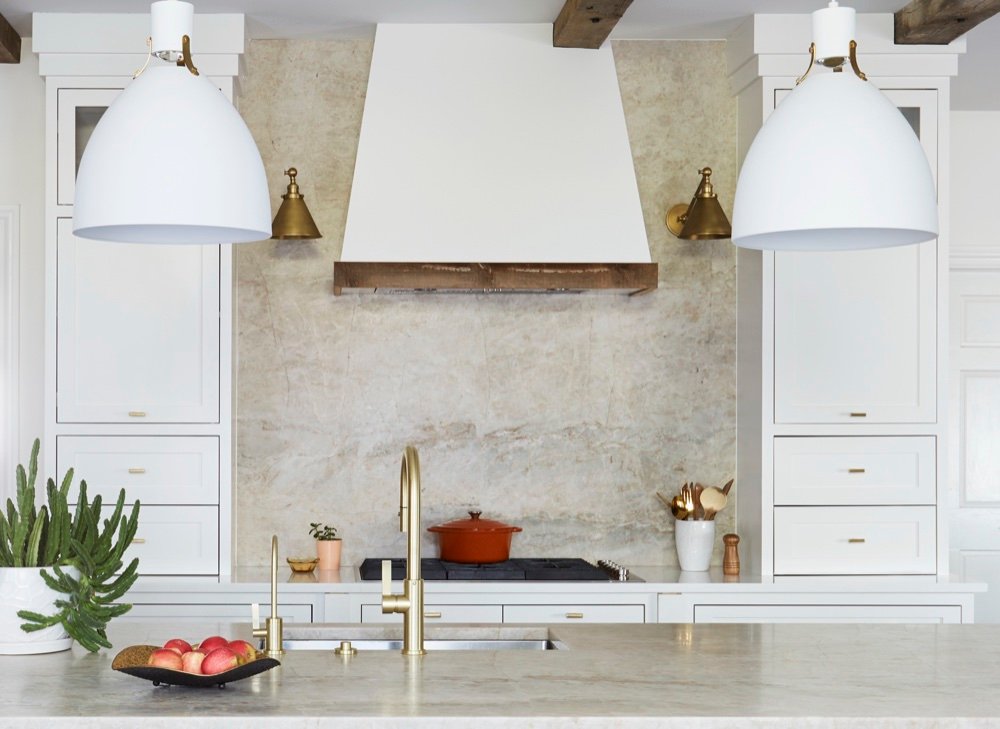 Symmetrical kitchen range wall with cinnamon-veined quartzite slab splash, white plaster hood banded in reclaimed wood, brass cone sconces, matte white pendants and flanking counter towers, by Martire Custom Homes