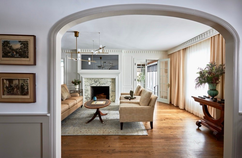 View through an arched opening into a living room with a veined marble fireplace, brass chandelier, and antique framed prints, by Martire Custom Homes