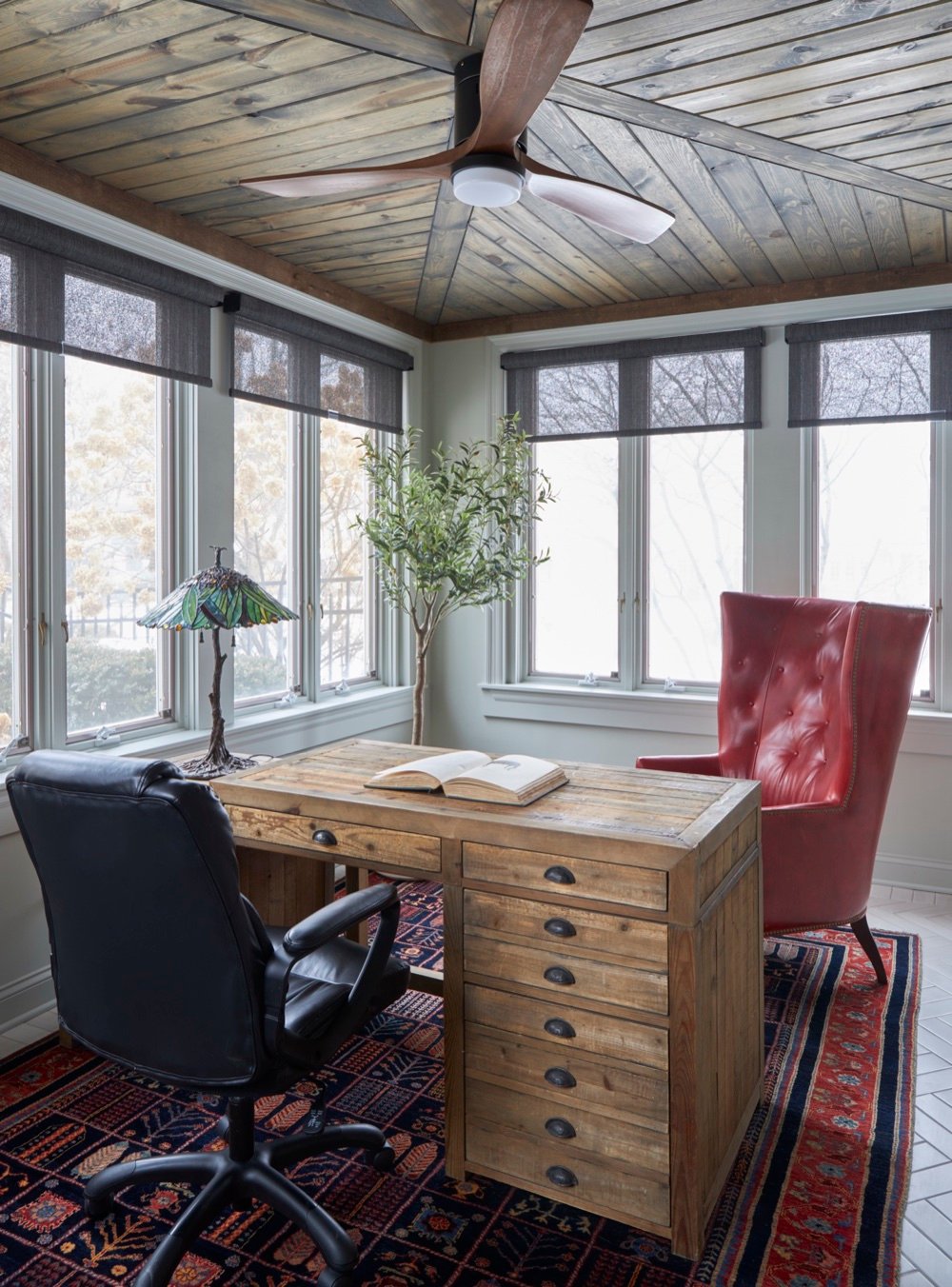 Sunlit gentleman's office with rustic reclaimed-wood desk, red tufted leather wing chair, stained-glass lamp, and wood plank ceiling, by Martire Custom Homes