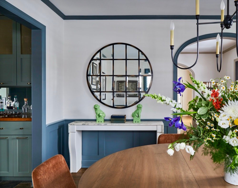 Dining room with slate-blue wainscoting, round gridded mirror over a marble-topped console, and rust velvet chairs, by Martire Custom Homes