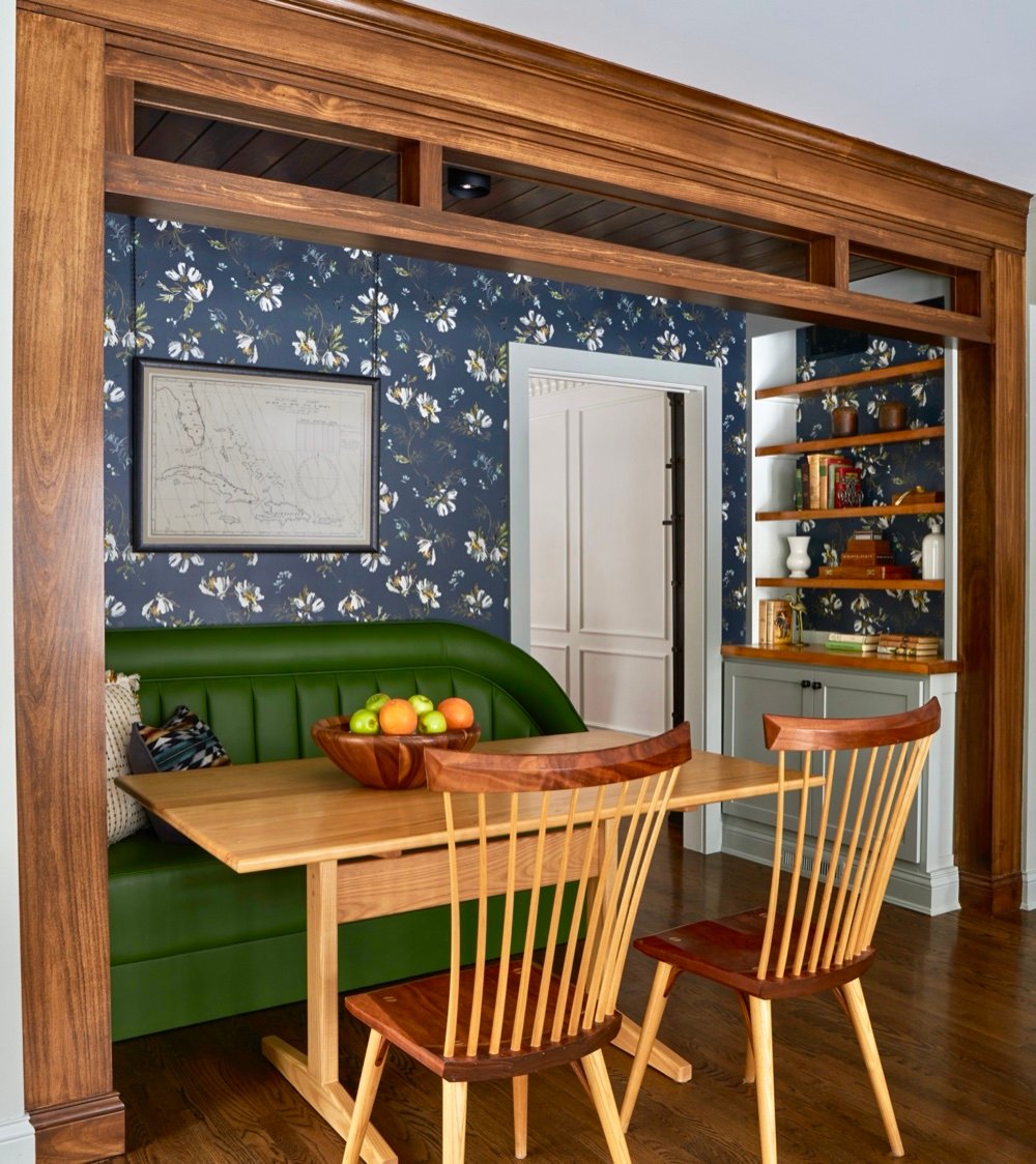 Study nook with channel-stitched green leather banquette, pale wood trestle table, spindle-back chairs, navy floral wallpaper, and stained millwork surround with transom windows, by Martire Custom Homes