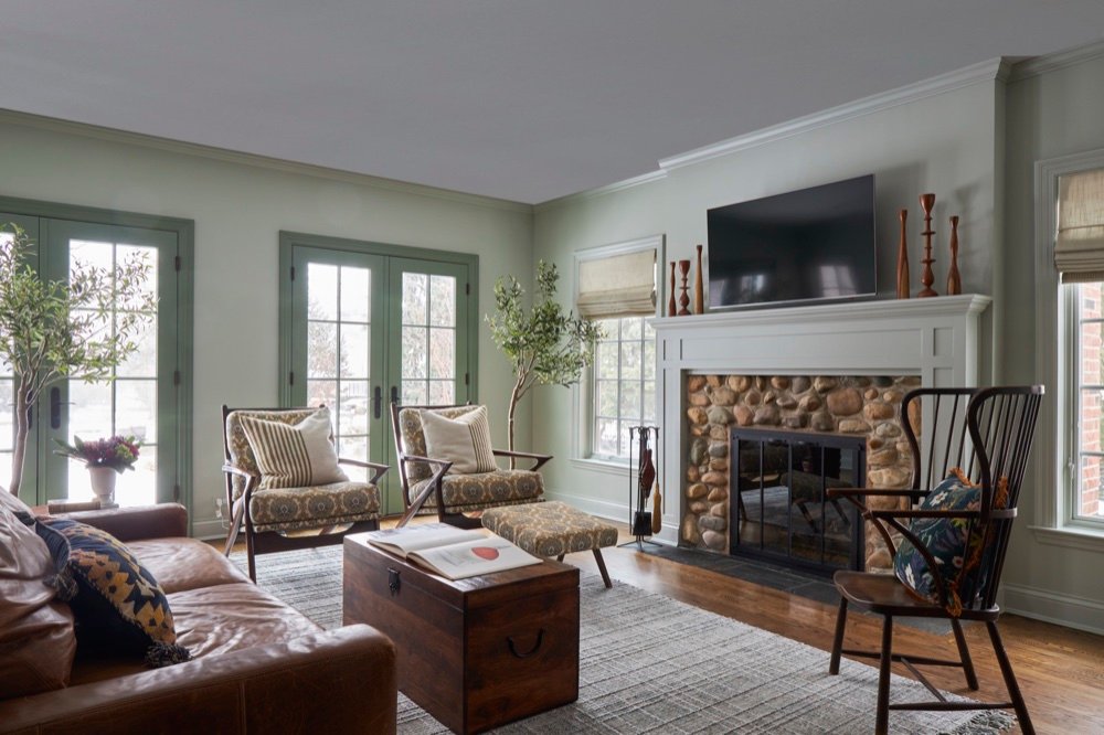 Family room with river-rock stone fireplace, sage-green French doors, patterned mid-century armchairs, and cognac leather sofa, by Martire Custom Homes