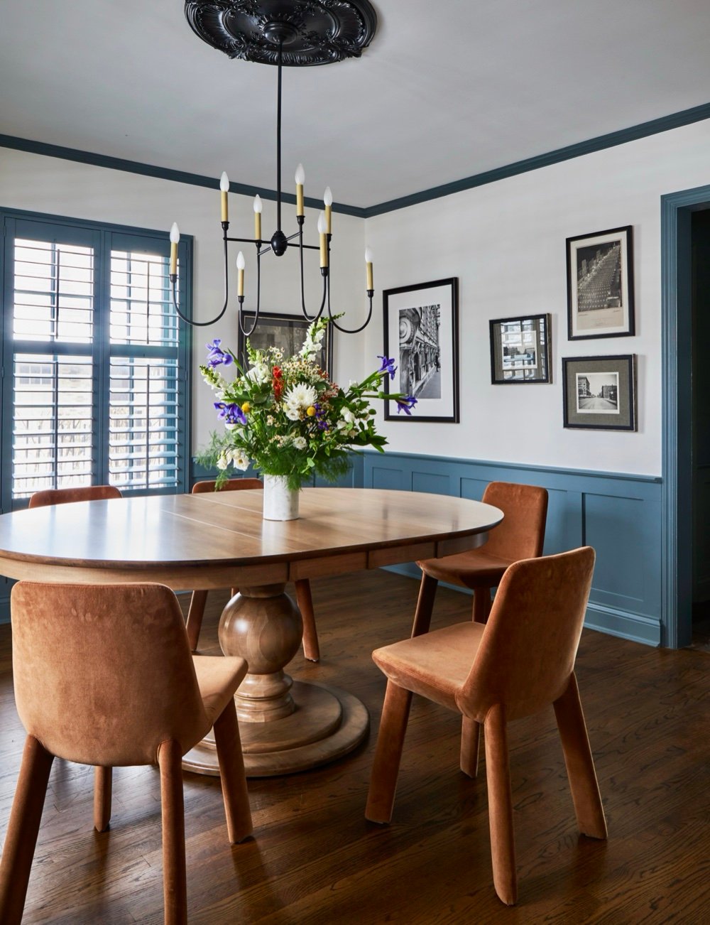 Dining room with round pedestal table, rust velvet chairs, blue wainscoting and shutters, black chandelier on ornate medallion, and gallery of framed photographs, by Martire Custom Homes