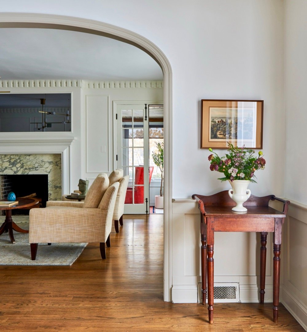 Arched opening from the hall into the living room, with an antique turned-leg washstand, white urn of flowers, and framed landscape print, by Martire Custom Homes