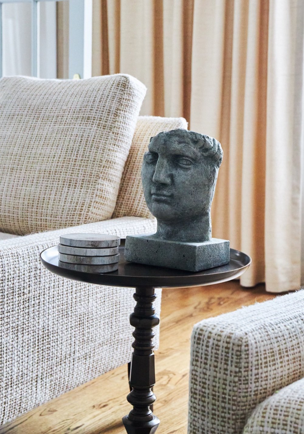Carved Roman head sculpture on a black pedestal table beside a cream armchair and drapery, by Martire Custom Homes