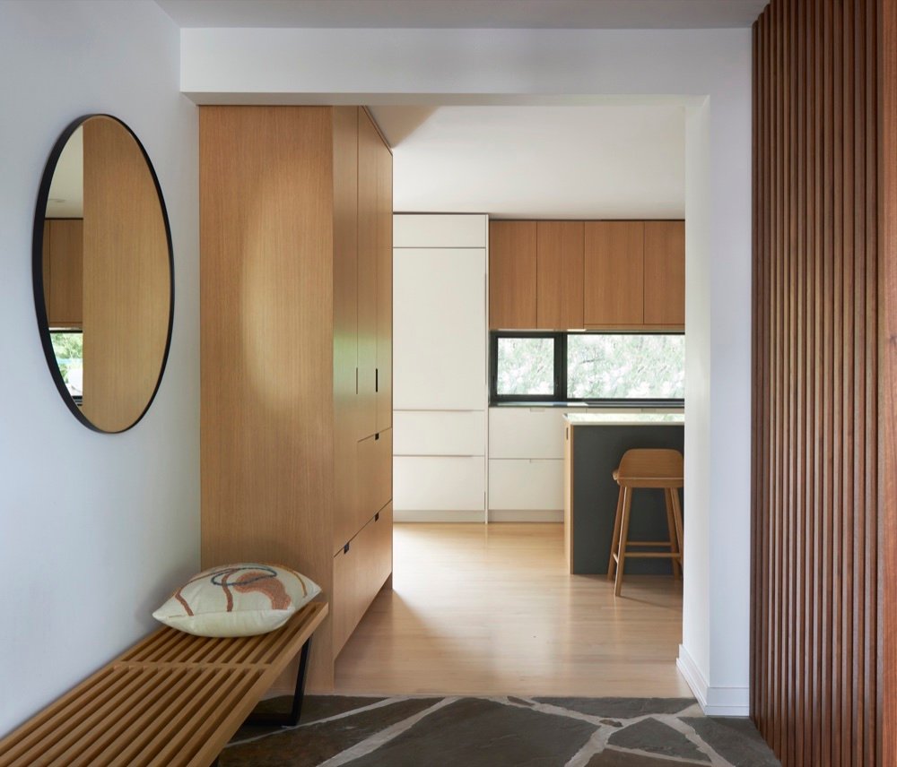 Entry foyer with a black-framed round mirror, white oak wardrobe cabinetry, slatted wood bench and flagstone floor opening to a Scandinavian-inspired kitchen by Martire Custom Homes