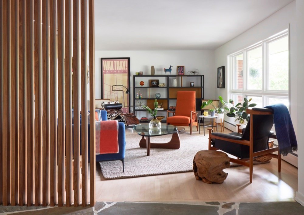 Living room with mid-century furnishings and large windows, seen past a walnut slat trim wall rising from the flagstone entry, by Martire Custom Homes