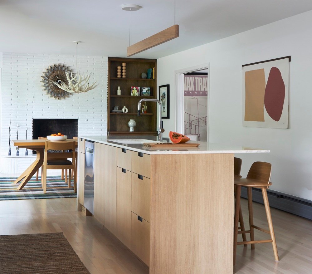 Scandinavian-inspired white oak kitchen island with marble counter and recessed pulls, opening to a dining room with white painted brick fireplace and walnut built-ins, by Martire Custom Homes
