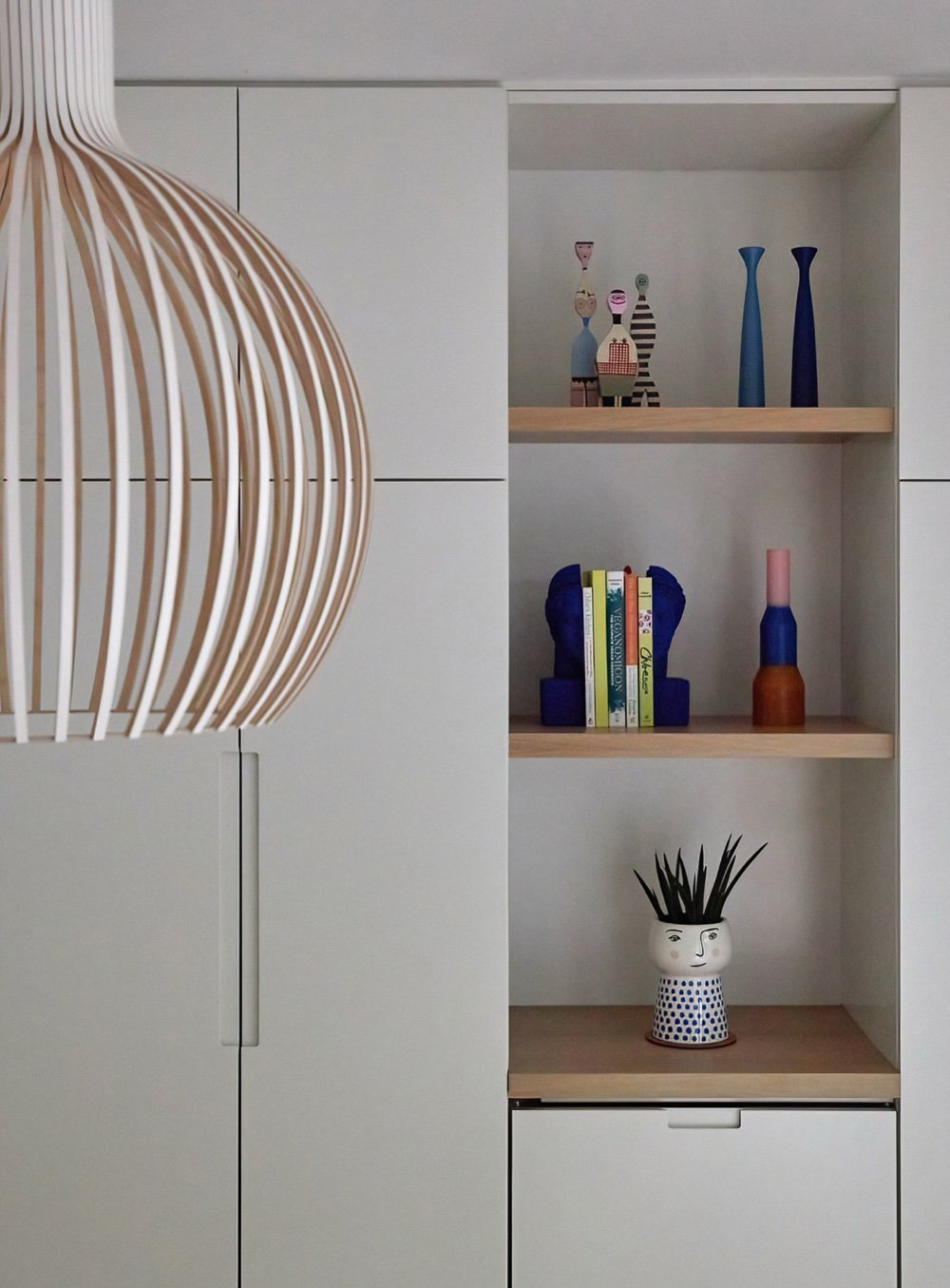 Detail of a slatted birch pendant beside floor-to-ceiling white cabinetry, its open oak shelves styled with folk figures, vases and cookbooks, by Martire Custom Homes