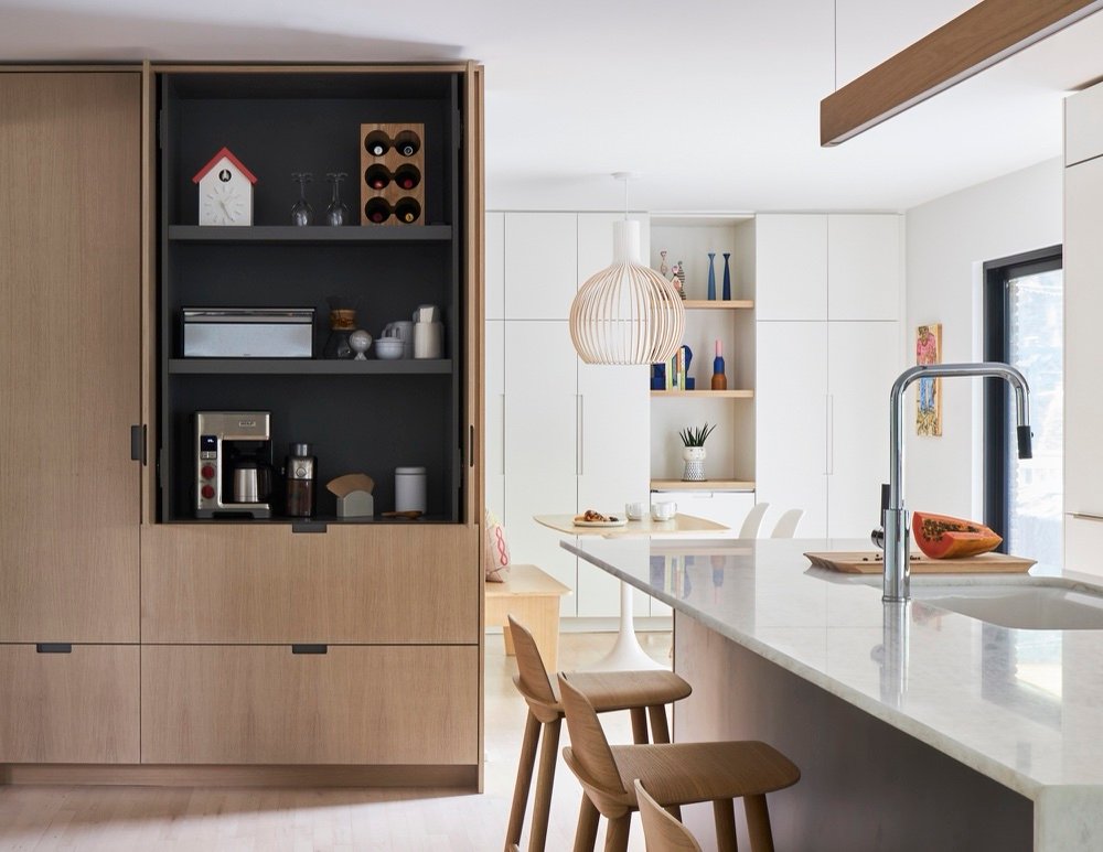 White oak pantry cabinet with pocket doors open to a charcoal-lined coffee station, with the marble island and dinette beyond by Martire Custom Homes