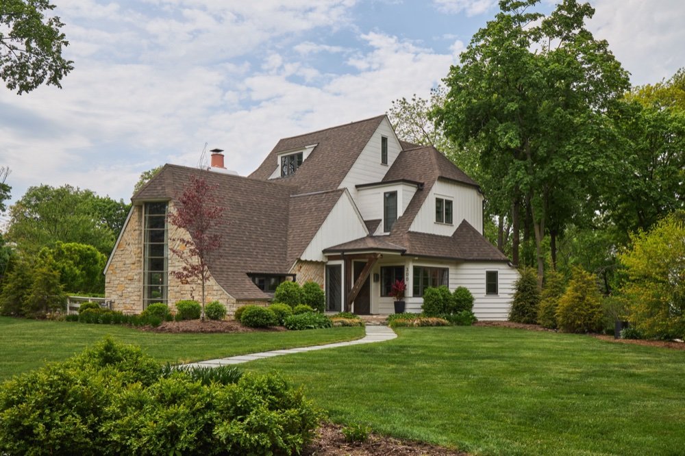 Front exterior of a restored 1940s storybook residence with a sweeping catslide roofline, limestone bay with vertical window band and white lap siding, by Martire Custom Homes