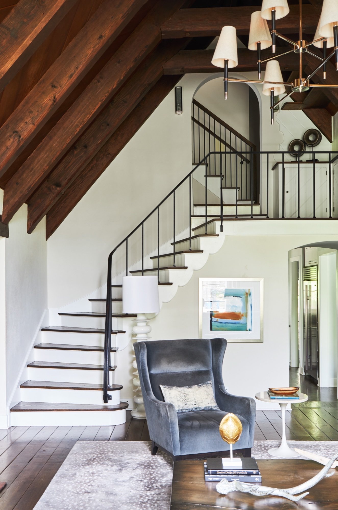 Curved staircase with slender iron railing and scalloped stringer rising to a loft balcony beneath restored dark timber rafters, with arched doorways beyond, by Martire Custom Homes