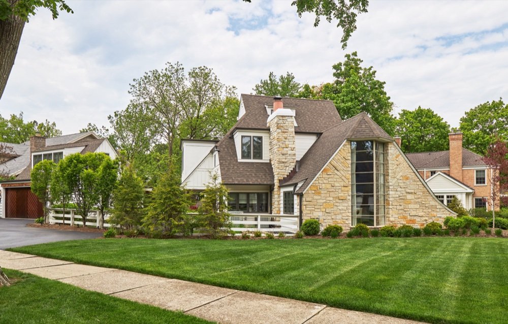 Side exterior of a restored storybook residence showing the limestone chimney, curved catslide eave and stone bay with vertical glazing, by Martire Custom Homes