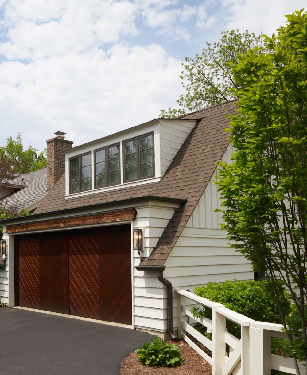 Detached garage with chevron-paneled wood doors, reclaimed timber header, shed dormer and brick chimney beyond, by Martire Custom Homes