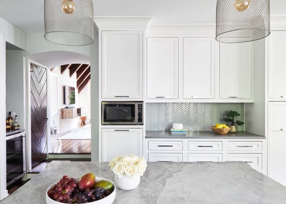 Kitchen with white inset cabinetry, built-in microwave and pale herringbone tile backsplash seen from the marble-topped island, a chevron-planked door standing open in an arched passage to the beamed great room, by Martire Custom Homes