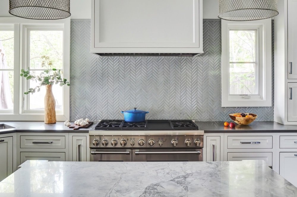 Professional-style stainless range and double ovens under a painted paneled hood, with glazed herringbone tile backsplash flanked by casement windows, by Martire Custom Homes