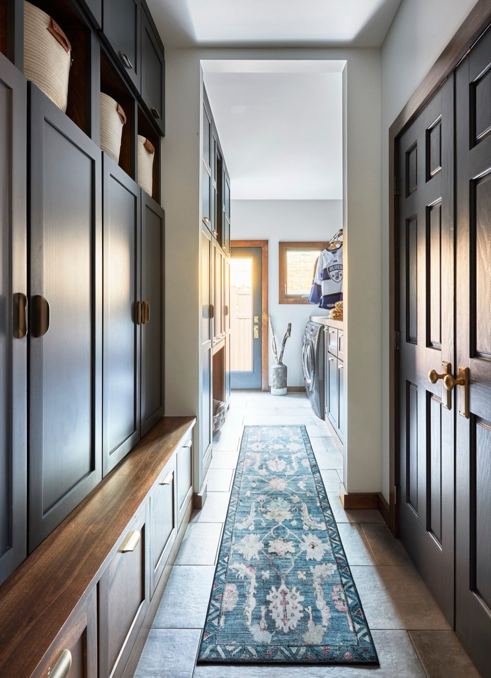 Mudroom corridor with charcoal painted lockers, walnut bench and drawers, and a blue patterned runner leading toward the sunlit laundry room, by Martire Custom Homes