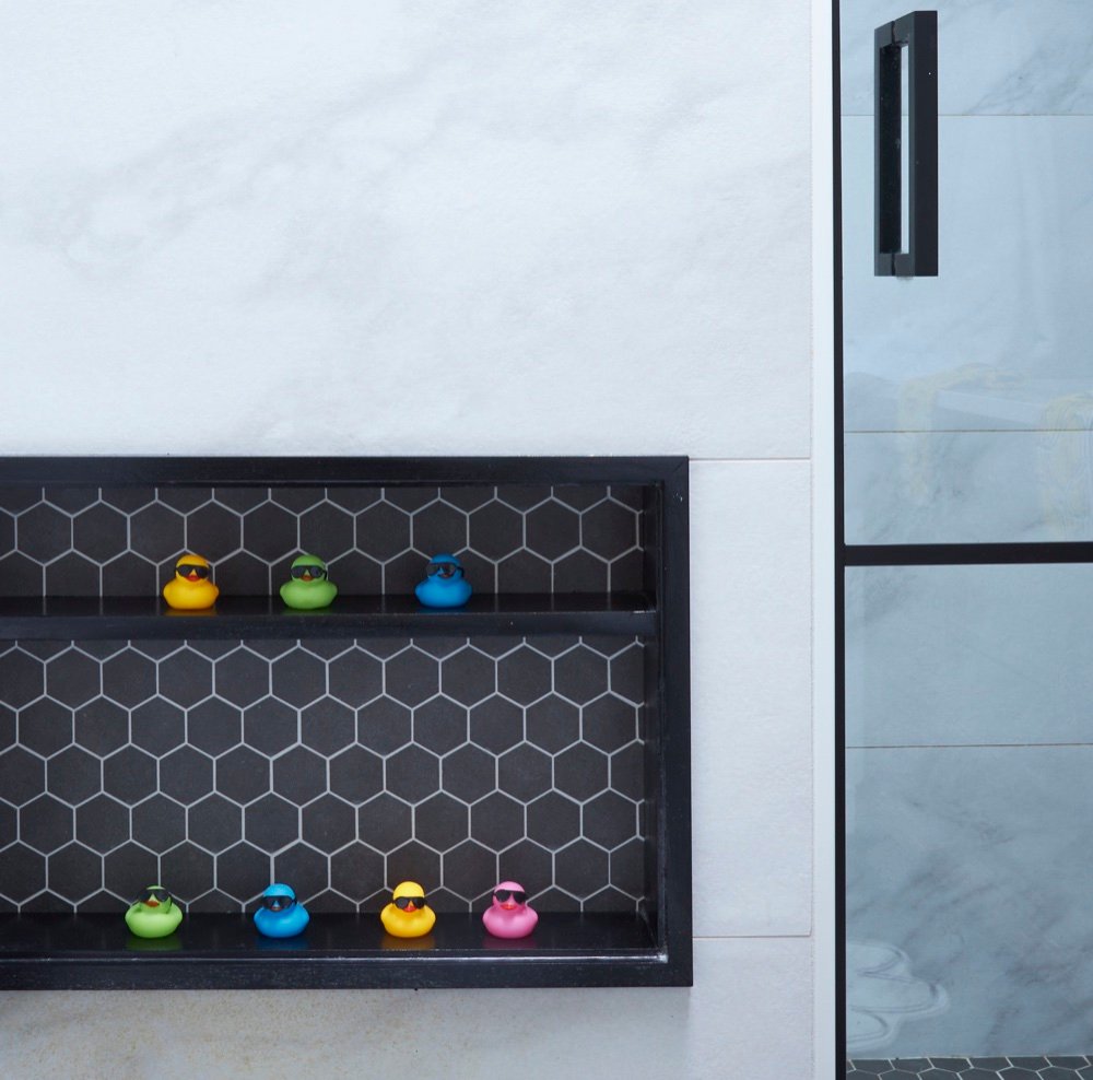 Black-framed shower niche lined with dark hexagon mosaic tile displaying colorful rubber ducks beside a black-framed glass shower door, by Martire Custom Homes