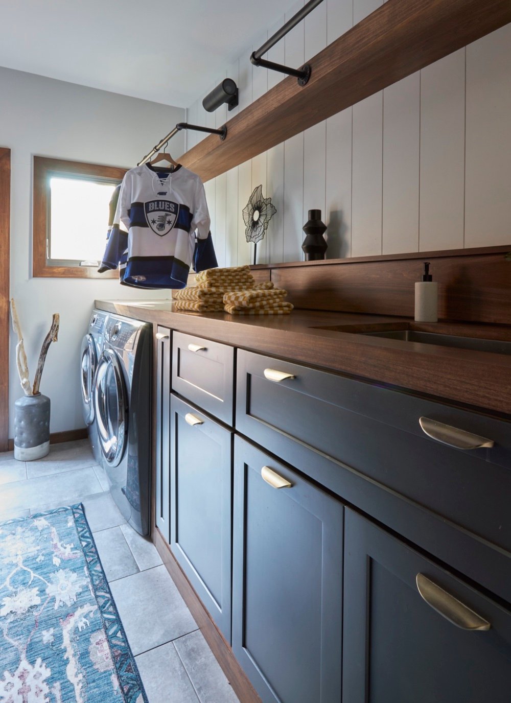 Laundry room run of charcoal cabinetry with brass pulls, walnut countertop and backsplash, shiplap walls and a blackened-pipe drying rail, by Martire Custom Homes