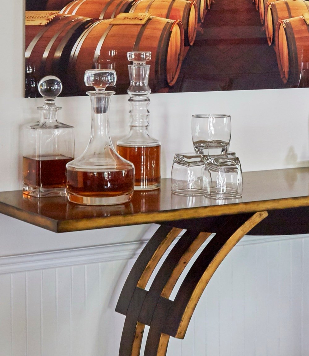 Bar vignette with crystal decanters and tumblers arranged on a gilt iron console against white beadboard wainscoting, by Martire Custom Homes