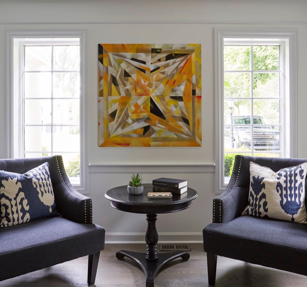 Sitting area with two navy nailhead-trimmed chairs flanking a black pedestal table beneath abstract artwork between tall windows, by Martire Custom Homes