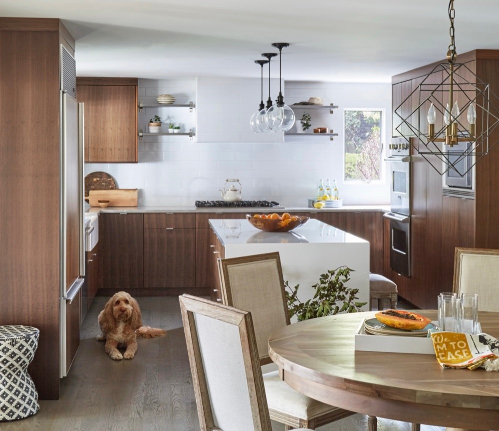 Open-plan kitchen and dinette with rift-cut walnut cabinetry, a white quartz waterfall island, round wood dining table and the family dog resting on grey oak floors, by Martire Custom Homes