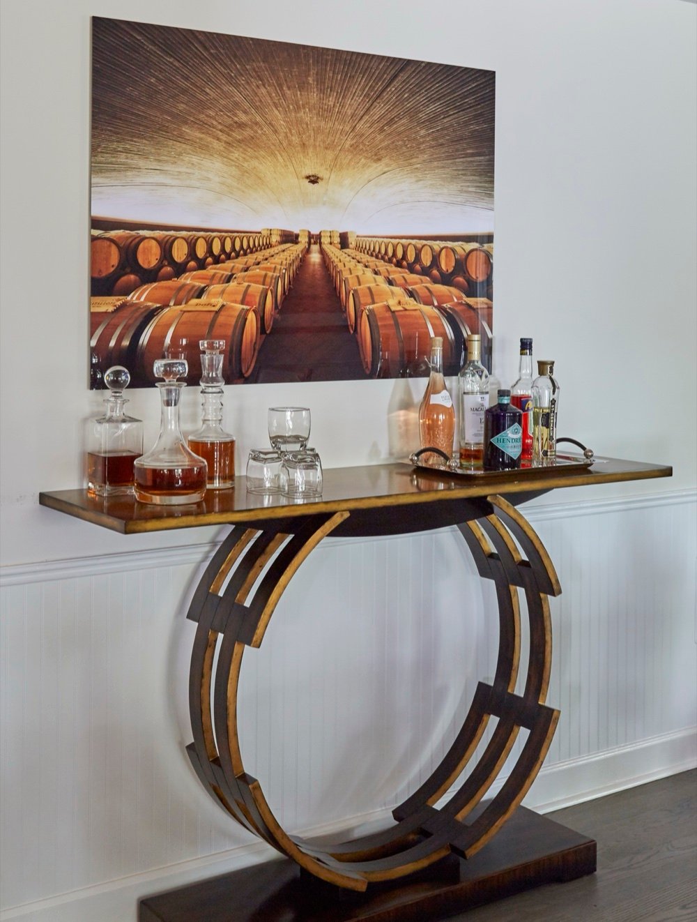 Bar console with a sculptural gilt ring base holding decanters and glassware beneath a large wine cellar photograph, by Martire Custom Homes