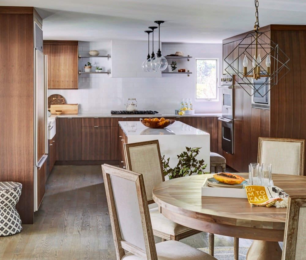 Dinette with linen-backed chairs and geometric brass chandelier opening onto a kitchen with walnut cabinetry and a white quartz waterfall island, by Martire Custom Homes