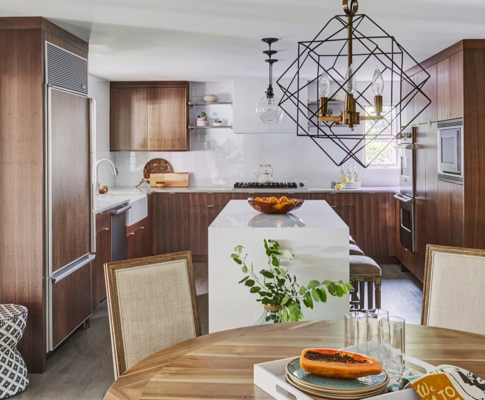 Kitchen seen past a geometric brass chandelier, with farmhouse sink, rift-cut walnut cabinetry and a white quartz waterfall island under glass pendants, by Martire Custom Homes