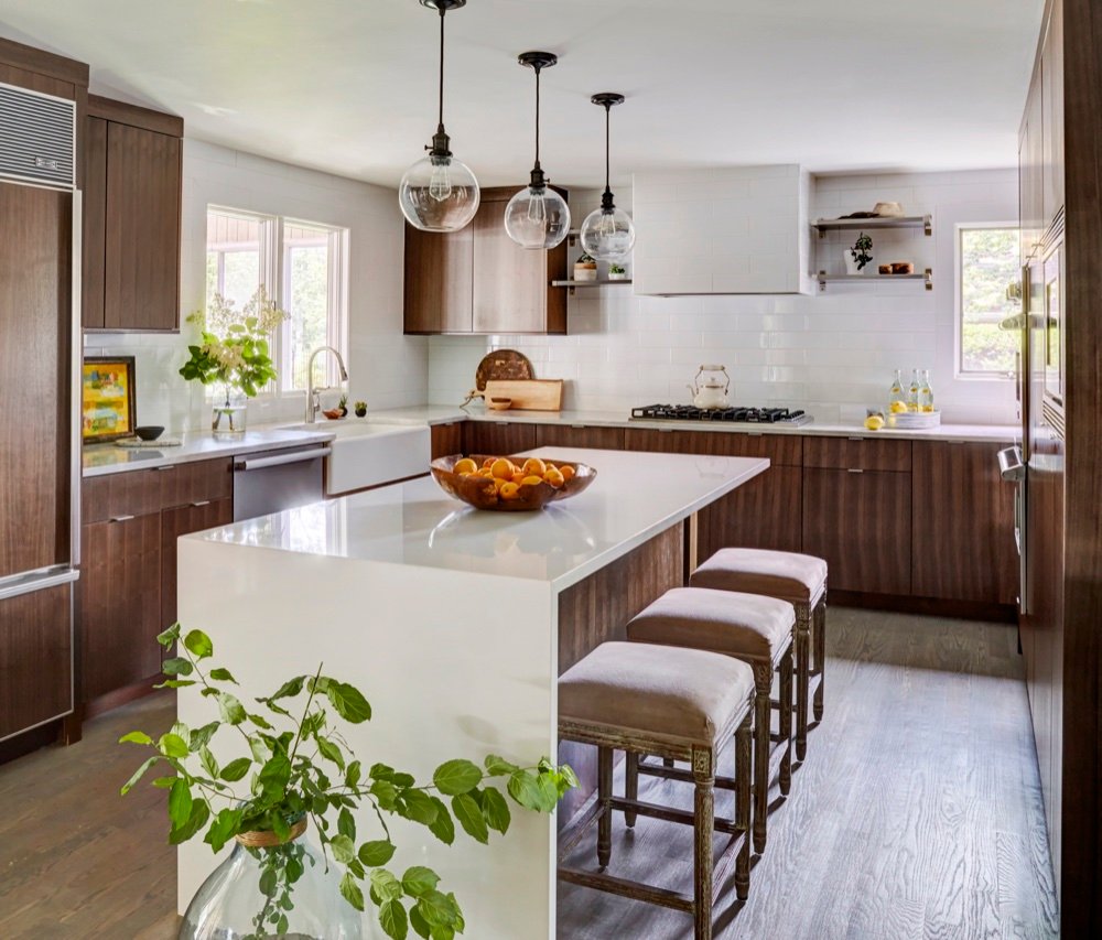 Renovated kitchen with a white quartz waterfall island, suede counter stools, rift-cut walnut cabinetry, farmhouse sink and white tiled hood, by Martire Custom Homes