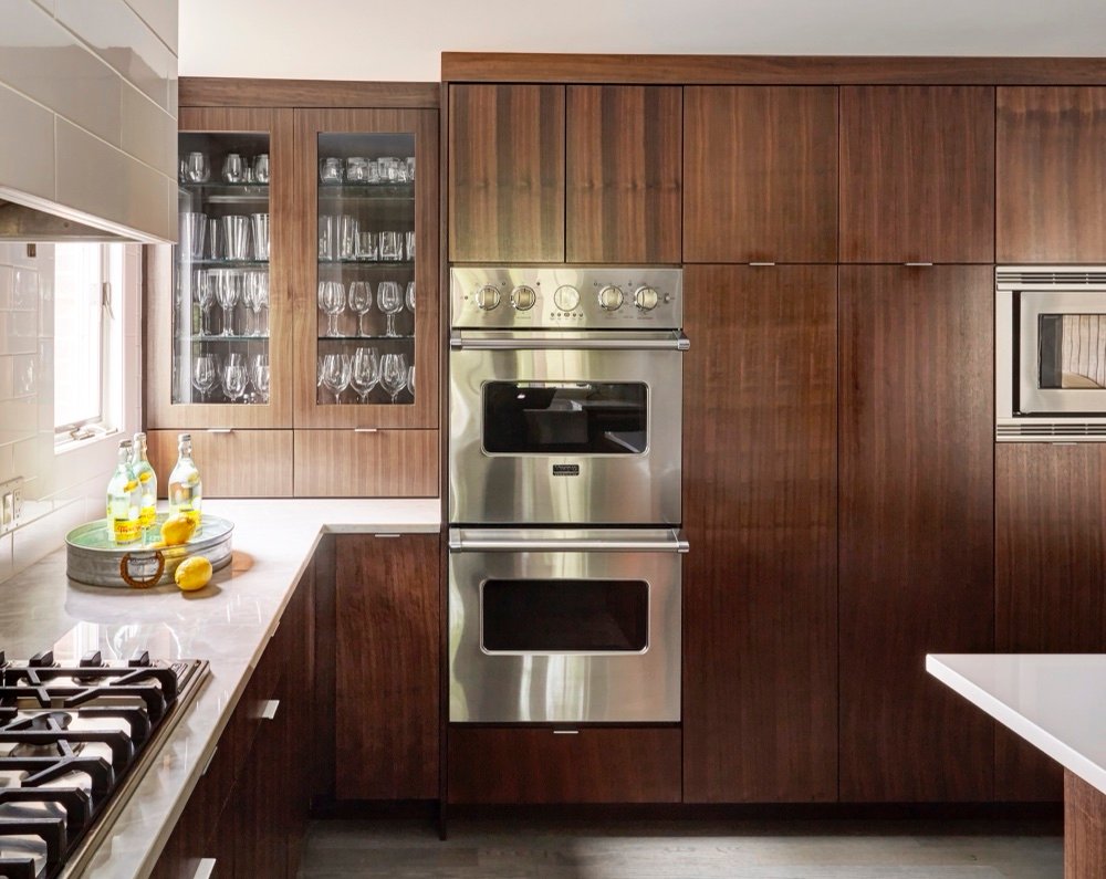 Floor-to-ceiling rift-cut walnut cabinetry with glass-front glassware display, stainless double wall ovens and built-in microwave, by Martire Custom Homes